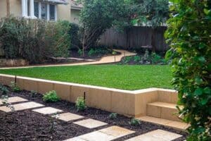 A smooth, light-coloured retaining wall bordering a manicured green lawn and mulched garden with square stepping stones.