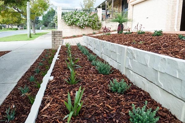 A low, two-tier Perth retaining wall built from cream, stone-textured blocks edging narrow, mulched residential garden beds planted with rows of small shrubs.